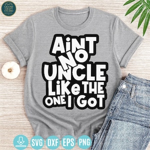 May include: A gray t-shirt with the text "Ain't No Uncle Like The One I Got" in a bold, white font with a black outline. The shirt is folded, with a pair of blue jeans visible on the right side. The shirt has rolled-up sleeves.