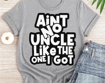 May include: A gray t-shirt with the text "Ain't No Uncle Like The One I Got" in a bold, white font with a black outline. The shirt is folded, with a pair of blue jeans visible on the right side. The shirt has rolled-up sleeves.