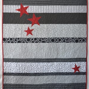 May include: A handmade quilt with a striped pattern in shades of grey and black, featuring three red star appliqués. The quilt has a red border and is likely made of cotton or a similar fabric.