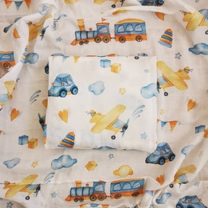 May include: White baby blanket with a repeating pattern of blue cars, yellow airplanes, trains, clouds, stars, and orange hearts. The blanket is folded and placed on top of a larger blanket with the same design.