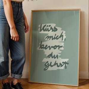 Puede incluir: Un p&oacute;ster enmarcado con el texto alem&aacute;n "K&uuml;ss mich, bevor du gehst" en escritura gris oscuro sobre un fondo verde azulado claro. El p&oacute;ster tiene un marco de madera delgado y marr&oacute;n claro y est&aacute; apoyado contra una pared.