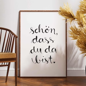 Könnte beinhalten: Ein gerahmter Kunstdruck mit dem deutschen Satz "schön, dass du da bist" in eleganter schwarzer Schrift auf weißem Hintergrund. Der Holzrahmen ergänzt die warmen Töne des Raumes, mit einem Stuhl und einer dekorativen Pflanze im Hintergrund.