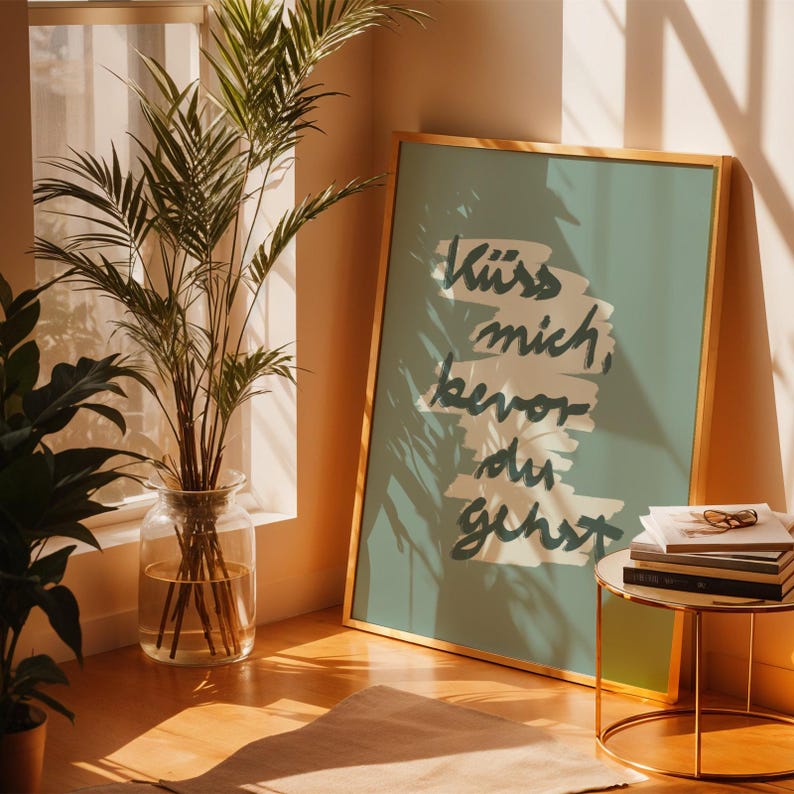 Puede incluir: Un p&oacute;ster enmarcado con el texto "K&uuml;ss mich, bevor du gehst" en escritura azul oscuro sobre un fondo verde azulado. El p&oacute;ster est&aacute; en un marco dorado y se apoya contra una pared. Un jarr&oacute;n de cristal con una planta est&aacute; cerca de una ventana.