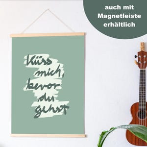 Puede incluir: Un estampado de arte mural verde salvia con la frase alemana "K&uuml;ss mich, bevor du gehst" en un estilo manuscrito. La impresi&oacute;n se sujeta con barras de madera y una cuerda para colgar. Tambi&eacute;n es visible el texto "auch mit Magnetleiste erh&auml;ltlich".