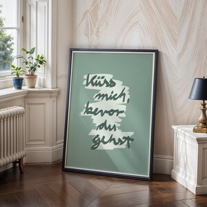 Puede incluir: Una impresi&oacute;n art&iacute;stica enmarcada con la frase alemana "K&uuml;ss mich, bevor du gehst" en escritura verde oscuro sobre un fondo verde salvia. La impresi&oacute;n est&aacute; en un marco negro y se apoya contra una pared.