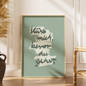 Puede incluir: Una impresi&oacute;n art&iacute;stica enmarcada con el texto "K&uuml;ss mich, bevor du gehst" en un estilo manuscrito. El texto es de color verde oscuro sobre un fondo verde claro. El marco es de color madera clara.
