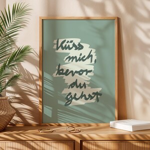 Puede incluir: Un cuadro enmarcado con la frase alemana "K&uuml;ss mich, bevor du gehst" en escritura azul oscuro sobre un fondo verde azulado. El marco de madera est&aacute; sobre una superficie de madera con anillos dorados.