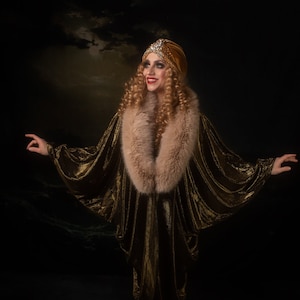 Peut inclure: Une femme portant une robe en velours or avec un col en fourrure et un turban orné de bijoux. Elle se tient sur un fond sombre et sourit.
