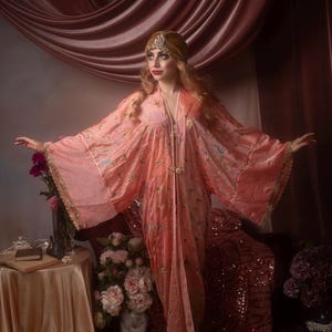 Puede incluir: Una bata fluida de color melocotón con mangas anchas y bordados florales. La bata presenta un cierre y ribete dorados. El fondo incluye una cortina rosa, flores y una pequeña mesa. La estética general es vintage y elegante.