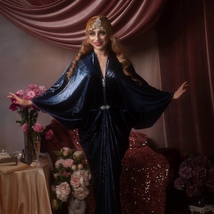 May include: A woman in a dark blue, flowing velvet dress with wide sleeves and a jeweled belt. She wears a gold headpiece and stands in front of a pink curtain, with flowers and a small table to the side. The dress has a V-neck.