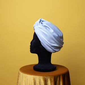Może przedstawiać: Jasnoniebieski turban wykonany z miękkiej tkaniny. Turban jest prezentowany na czarnej głowie manekina na żółtym tle.
