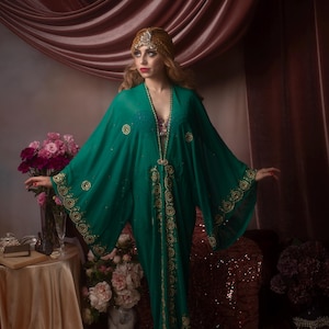 May include: A flowing emerald green kaftan with gold embroidery and embellishments. The kaftan features wide sleeves and a V-neckline. A gold headpiece and necklace complement the garment. The background includes pink and maroon drapery and a table with flowers.