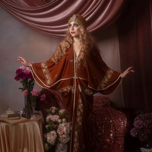 May include: A woman in a rust-colored, flowing gown with gold embroidery and a matching headpiece. The dress has wide sleeves and a cinched waist. The background features pink drapes and floral arrangements, creating a vintage, bohemian aesthetic.