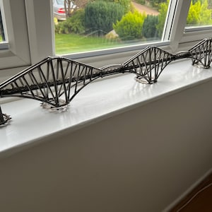 May include: A metal sculpture of the Forth Rail Bridge, a famous landmark in Scotland. The sculpture is made of thin metal rods and is painted in a silver colour. It is displayed on a white windowsill.