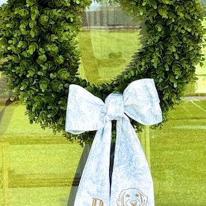 May include: A green artificial boxwood wreath with a blue and white patterned bow. The bow has a gold letter "D" and a golden retriever dog embroidered on it.