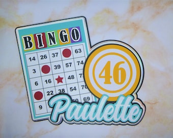 May include: A blue and yellow bingo card with the word "BINGO" in colorful letters. The card has numbers and a red dot, a red circle, and a red star. A yellow and orange bingo chip with the number "46" is next to the card. The word "Paulette" is written in blue and white letters below the chip.