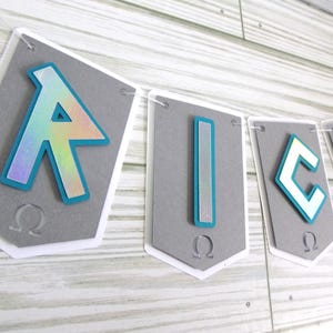 May include: A banner spelling out the word "AICK" with gray pennants. Each pennant has a white border and a gray background. The letters are in a teal outline with a holographic, rainbow-colored fill. The banner is strung on a white string.