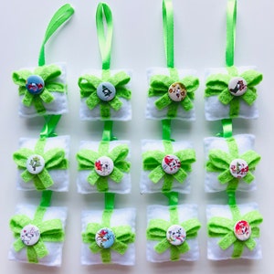 May include: Twelve green and white felt Christmas ornaments shaped like presents with a bow and a button on the front. Each button has a different Christmas-themed design.
