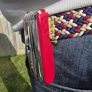 May include: A red multi-tool knife clipped to a blue denim pocket. The knife has multiple silver tools and a red handle. The person is wearing a woven belt with blue, red, and tan colors.