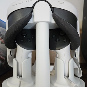 Könnte beinhalten: Weißer VR-Headset-Ständer mit zwei Controllern. Das Headset hat ein schwarzes Stirnband und die Controller haben schwarze Knöpfe. Der Ständer hat eine weiße Basis und eine weiße Stange.