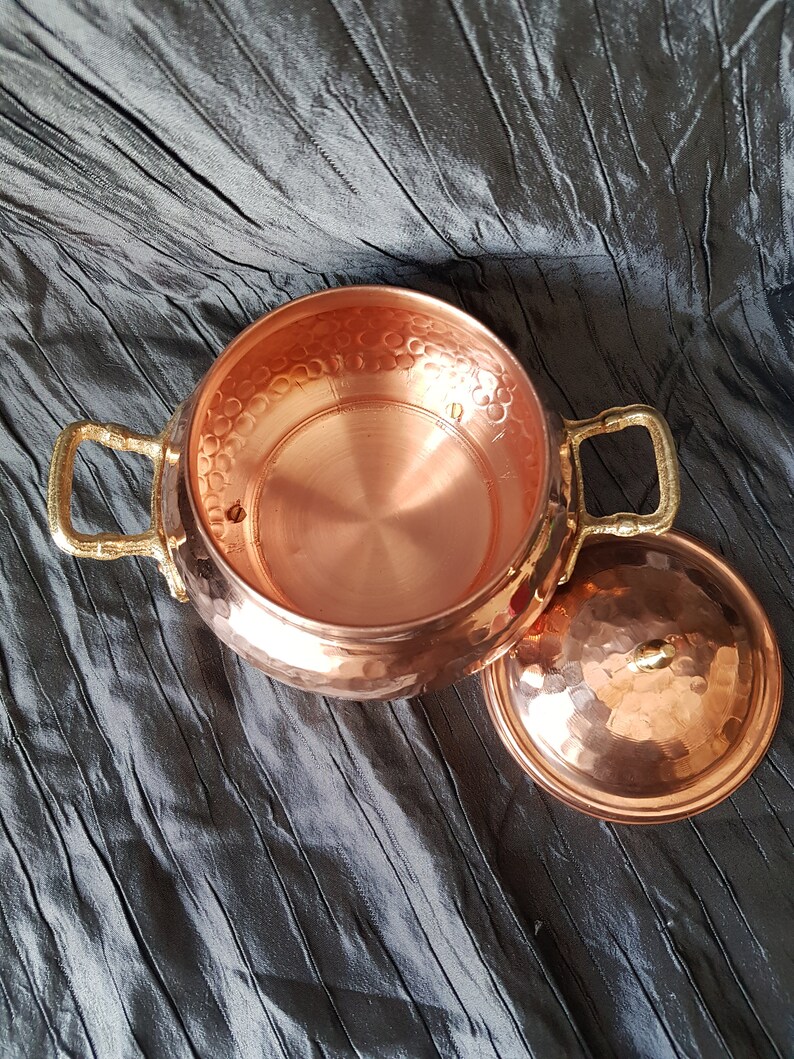 Puede incluir: Una olla de cobre con un acabado martillado y dos asas de lat&oacute;n. La olla tiene una tapa con un acabado martillado similar.