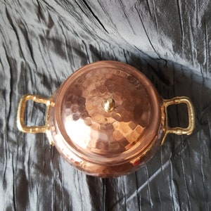 Puede incluir: Una olla de cobre con acabado martillado y dos asas doradas. La olla tiene una tapa abovedada con un peque&ntilde;o pomo en la parte superior.