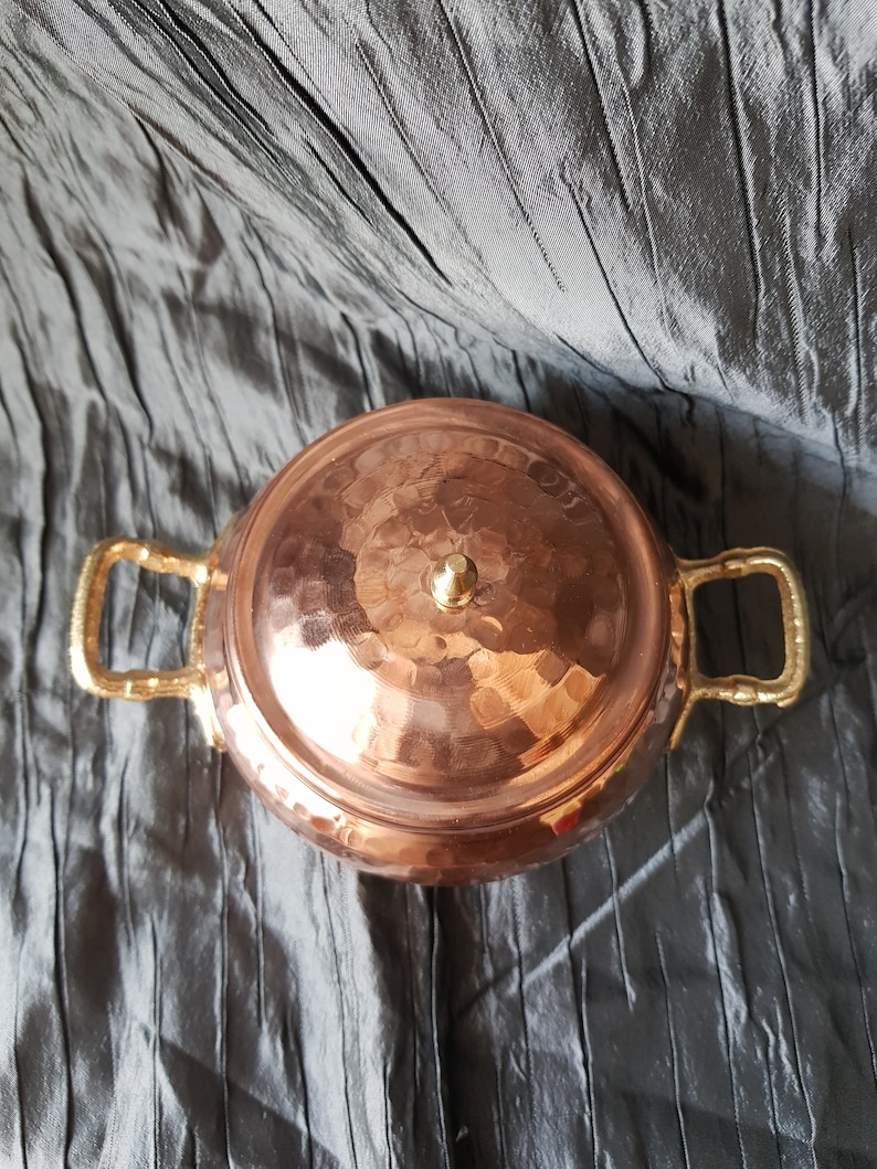 Puede incluir: Una olla de cobre con acabado martillado y dos asas doradas. La olla tiene una tapa abovedada con un peque&ntilde;o pomo en la parte superior.