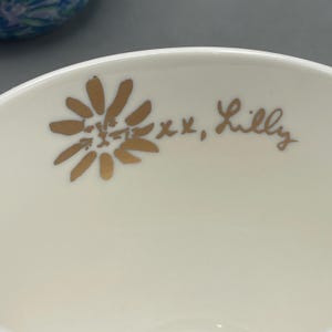 Puede incluir: Primer plano de un cuenco de cer&aacute;mica color crema con un dise&ntilde;o floral dorado y el texto manuscrito "xx, Lilly". El dise&ntilde;o floral es una margarita estilizada.