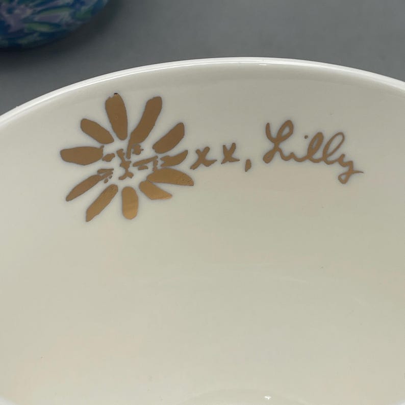 Puede incluir: Primer plano de un cuenco de cer&aacute;mica color crema con un dise&ntilde;o floral dorado y el texto manuscrito "xx, Lilly". El dise&ntilde;o floral es una margarita estilizada.