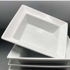 Puede incluir: Una pila de fuentes rectangulares blancas de cerámica. Cada fuente tiene un centro ligeramente hundido y un diseño limpio y moderno. Las fuentes están apiladas, mostrando su forma.