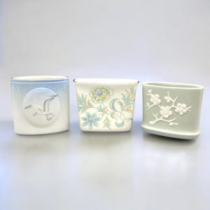 May include: Three decorative ceramic containers. One is light blue with a bird design, another is white with floral patterns, and the third is pale green with a floral relief. Each has a unique shape and design, suitable for small plants or decor.