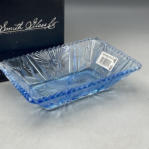 May include: A rectangular, light blue glass dish with a starburst and leaf pattern. The dish has a scalloped edge and is labeled "Smith Glass Co." The dish is approximately 8 inches long and 5 inches wide.