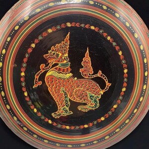 May include: A round, black decorative plate with a detailed, colorful design. The plate features a central image of a mythical creature in red, yellow, and green, surrounded by concentric rings of color and dotted patterns. The plate has a glossy finish.