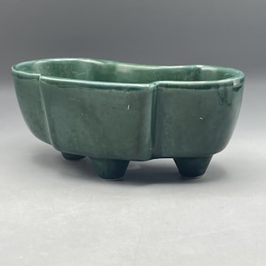 May include: A dark green ceramic planter with an irregular oval shape and three small feet. The planter is glazed and has a smooth finish.