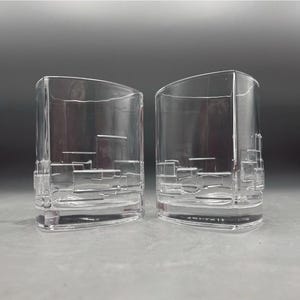 Peut inclure: Deux verres à boire rectangulaires transparents avec un dessus incurvé. Les verres présentent un motif géométrique décoratif gravé dans le verre. Les verres sont en verre transparent.