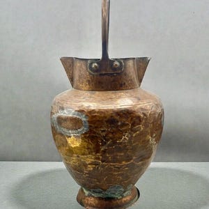 May include: A hammered copper pitcher with a wide, rounded body, a flared rim, and a tall, arched metal handle. The surface features a textured, dimpled finish with visible variations in color, including dark brown, bronze, and patches of green patina.