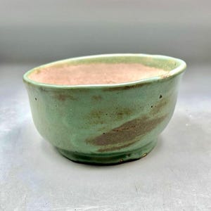 May include: A handmade ceramic bowl with a light green exterior and a pinkish-brown interior. The bowl has a rounded shape with a slightly angled rim. The exterior features brown streaks, adding a rustic touch. This bowl is suitable for serving food or as a decorative item.