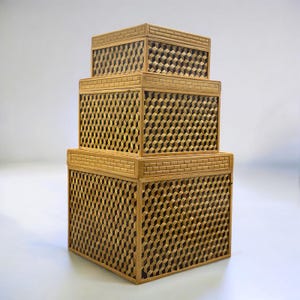 May include: Three wooden nesting boxes with a geometric cube pattern. The boxes are stacked, with the largest at the bottom and the smallest on top. The boxes have a light brown color and a brick-like border.