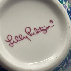 Puede incluir: Primer plano del interior de un cuenco de cer&aacute;mica blanca con el texto cursivo "Lilly Pulitzer" en magenta. El borde del cuenco presenta un colorido estampado floral en tonos azules, verdes y morados.