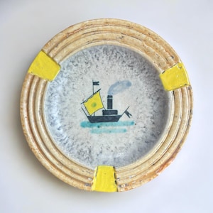 May include: Round ceramic plate with a cream-colored, ridged border and yellow accents. The center features a painted image of a black boat with a yellow sail, blue water, and a smokestack emitting blue smoke.