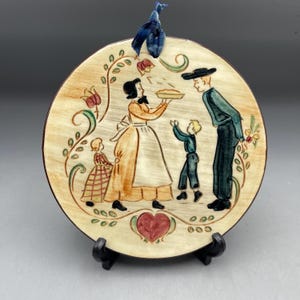 May include: A round ceramic decorative plate featuring a family scene with a woman holding a pie, a man, and two children. The plate has a beige background with floral and heart accents, and a blue ribbon for hanging.