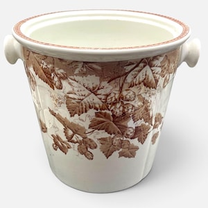 May include: A cream-colored ceramic wine cooler featuring a brown transferware pattern of hop vines and leaves. The vessel has two side handles and a decorative brown border around the rim, with a wide opening and a tapered base.