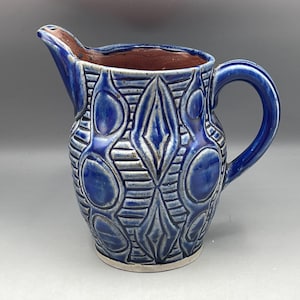 Puede incluir: Una jarra de cerámica azul y blanca con un patrón geométrico. La jarra tiene un cuerpo redondeado y un asa.