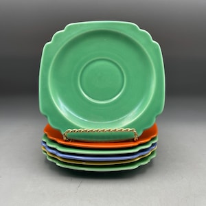 May include: A stack of five square ceramic saucers with scalloped edges. The top saucer is light green, followed by an orange saucer, a light green one, a light blue one, and a yellow one at the bottom. Each saucer features a central circular indentation.