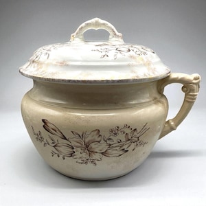 Op de afbeelding: Crèmekleurige antieke po met een deksel en handvat. De pot heeft een bloemenontwerp in bruintinten. Het deksel heeft een handvat en is versierd met vergelijkbare bloempatronen. De handgreep aan de zijkant is sierlijk.