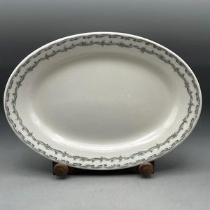 May include: An oval white ceramic serving platter featuring a decorative border pattern of grey floral garlands and ribbons. The dish has a smooth, glossy finish and is displayed on a small wooden stand, showcasing its classic shape and design.