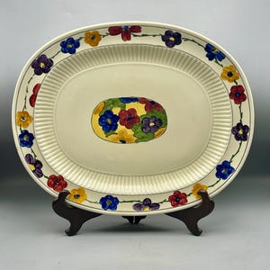 Può includere: Un piatto da portata ovale bianco con un motivo floreale. Il bordo presenta una ghirlanda continua di fiori rossi, gialli, blu e viola. Il centro del piatto presenta un gruppo di fiori più grande negli stessi colori.