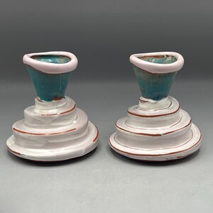 May include: Two ceramic candle holders. Each has a teal-colored top with a pink rim, atop a white, spiral-shaped base with reddish-brown accents. The candle holders are displayed against a gray background.