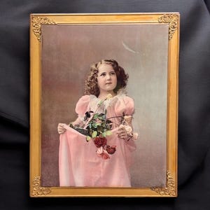 May include: A framed vintage portrait of a young girl in a pink dress holding roses. The gold frame has ornate corner details. The girl has curly hair and is looking upwards. The background is a soft, muted colour.