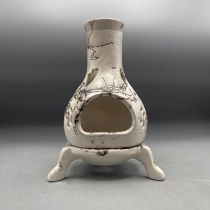 May include: A white ceramic chiminea with a bulbous body and a wide front opening. The surface features thin, dark, irregular black lines resembling smoke or crackle patterns. It stands on three curved legs and has small cutouts near the top.